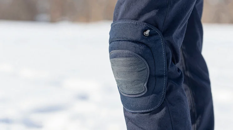 Close-up of articulated knee with reinforced stitching and gusset detail, snow visible in background